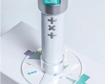 Könnte beinhalten: Ein weißer, zylindrischer Leuchtstabgriff mit einem silbernen, metallischen TXT-Logo, das vertikal in der Mitte ausgerichtet ist. Der Griff ist auf einem runden, transparenten Acrylsockel mit weißem Sternmuster und kleinen blauen Pluszeichen montiert.