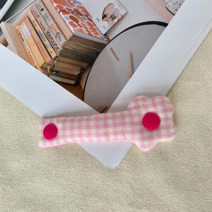 Puede incluir: Una pinza para el pelo de tela de cuadros rosa y blanco con forma de flor. La pinza tiene un bot&oacute;n rosa en el centro de la flor y otro en el extremo. La pinza est&aacute; sobre una superficie blanca.