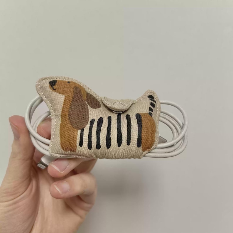 May include: A beige fabric cable tidy in the shape of a dachshund dog. The dog has brown ears and a brown patch on its back, with black stripes on its body. A white cable is wrapped around the tidy.