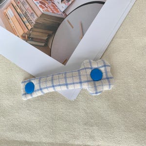 Puede incluir: Pinza para el pelo de tela a cuadros azules y blancos con dos botones circulares de color azul brillante. La pinza, con forma de hueso, est&aacute; sobre una superficie de color neutro.