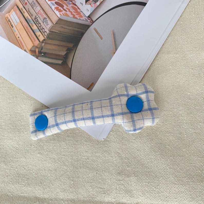 Puede incluir: Pinza para el pelo de tela a cuadros azules y blancos con dos botones circulares de color azul brillante. La pinza, con forma de hueso, est&aacute; sobre una superficie de color neutro.