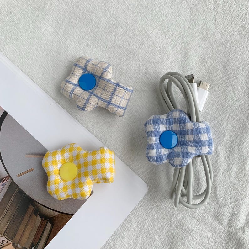 Puede incluir: Tres organizadores de cables de tela con forma de flor. Uno es a cuadros amarillos y blancos con un bot&oacute;n amarillo, otro es a cuadros azules y blancos con un bot&oacute;n azul, y el tercero es a cuadros azules y blancos con un bot&oacute;n azul. Sobre una superficie blanca.