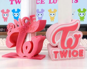 Könnte beinhalten: Zwei 3D-gedruckte Kunststoffständer für Leuchtstäbe. Der linke Ständer ist dunkelrosa mit dem Text ONCE und THIS IS FOR, während der rechte Ständer hellrosa ist und das TWICE-Logo sowie das Wort TWICE in Blockbuchstaben an der Basis zeigt.