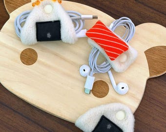 Könnte beinhalten: Drei plüschige Kabelorganizer in Form von Onigiri-Reisbällchen. Einer ist mit orangefarbenen Perlen verziert, einer hat einen Stoffaufsatz mit Lachsmuster und einer ist schlicht weiß mit einem schwarzen Stoffstreifen. Jeder hat einen weißen Knopf.