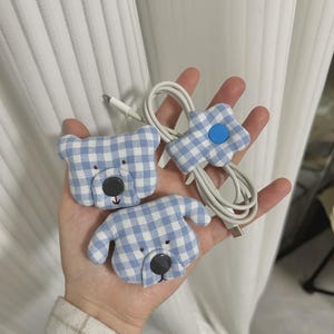 Puede incluir: Tres organizadores de cables con forma de oso, perro y flor. Los organizadores est&aacute;n hechos de tela de cuadros vichy azul y blanco con ojos y narices de bot&oacute;n negro. Un cable blanco es sujetado por el organizador en forma de flor.