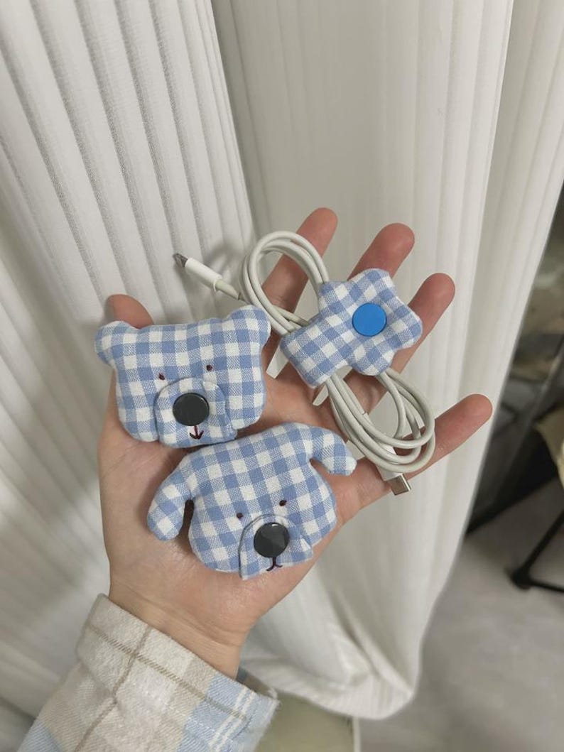 Puede incluir: Tres organizadores de cables con forma de oso, perro y flor. Los organizadores est&aacute;n hechos de tela de cuadros vichy azul y blanco con ojos y narices de bot&oacute;n negro. Un cable blanco es sujetado por el organizador en forma de flor.