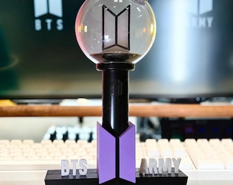 Könnte beinhalten: Ein BTS-Leuchtstab mit einer klaren, kugelförmigen Kappe, die ein schwarzes BTS-Logo enthält, montiert auf einem schwarzen zylindrischen Griff. Die Basis ist ein schwarzer rechteckiger Ständer mit einem lila BTS-Logo und weißem Text.