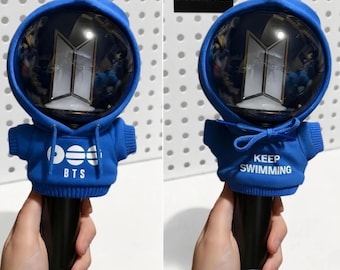 Könnte beinhalten: Ein schwarzer BTS-Leuchtstab in einem blauen Stoff-Hoodie-Überzug. Die Vorderseite des Hoodies zeigt den weißen Schriftzug 'BTS' mit einer kreisförmigen Grafik, während die Rückseite den weißen Text 'KEEP SWIMMING' und eine Kapuzenkordel aufweist.