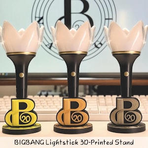 May include: Three lightstick stands featuring a crown-shaped top, a black handle with a gold ring, and a base shaped like the letter B with the number 20 inside. The stands are yellow, orange, and metallic gray. Text reads: BIGBANG Lightstick 3D-Printed Stand.
