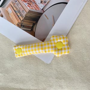 Puede incluir: Una pinza para el pelo de tela de cuadros amarillos y blancos con un extremo en forma de flor y dos botones amarillos. La pinza se encuentra sobre un fondo neutro, lo que sugiere un enfoque en los accesorios.