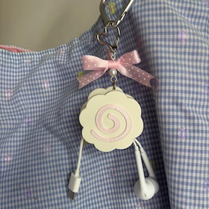 Può includere: Un organizzatore di cavi circolare bianco con bordi smerlati e un motivo a spirale rosa. È fissato a una chiusura argentata a forma di cuore e a un moschettone, decorato con un nastro rosa chiaro a pois e una piccola perlina bianca. Un cavo è collegato.
