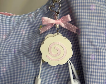 May include: A white, scalloped, circular cable organiser featuring a pink spiral design. It is attached to a silver heart-shaped clasp and a metal carabiner, accented with a light pink polka-dot ribbon and a small white bead. A white cable is attached.