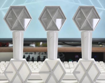 Könnte beinhalten: Drei weiße Leuchtstäbe mit einem sechseckigen Kopf und einem geometrischen EXO-Logo. Jeder Griff ist zylindrisch mit einem kleinen Logo-Emblem versehen, und die Basis ist ein weißer sechseckiger Ständer mit einem grauen Linienmuster.