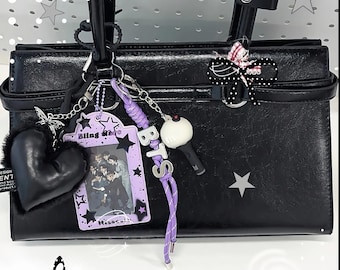 Könnte beinhalten: Eine schwarze Handtasche, verziert mit einem violetten Fotokartenhalter mit dem Text Bling stars, einem schwarzen Herzanhänger, einer violetten Kordel mit silbernen BTS-Buchstaben, einem kleinen weißen Leuchtstab-Anhänger und einer schwarz-weißen Schleife.