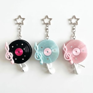Può includere: Tre portachiavi con design a disco in vinile circolare in nero, azzurro e rosa. Ognuno include una chiusura argentata a forma di stella, un ciondolo a chiave di violino, piccoli accenti a stella e auricolari bianchi con filo.