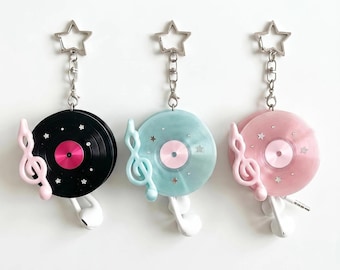 Könnte beinhalten: Drei Schlüsselanhänger mit kreisförmigen Schallplatten-Designs in Schwarz, Hellblau und Pink. Jeder Anhänger verfügt über einen silbernen sternförmigen Verschluss, einen Notenschlüssel-Anhänger, kleine Sternakzente und weiße Ohrhörer.