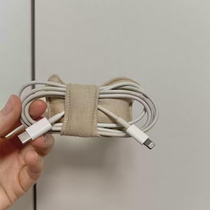 May include: A beige fabric cable tidy holding a white charging cable. The cable is coiled and secured with a matching fabric strap. The organiser is designed to keep cables neat and tidy.
