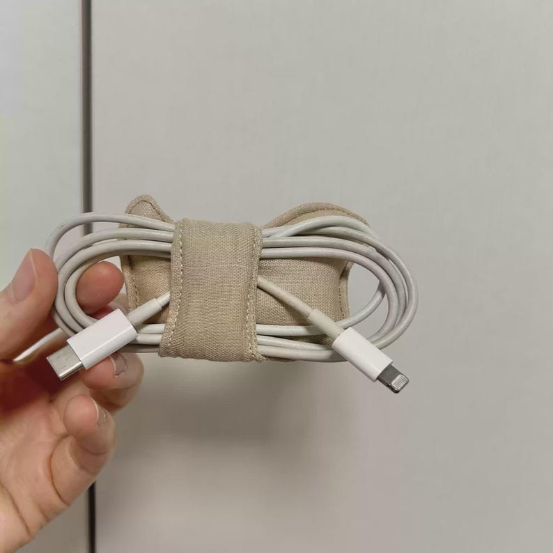 May include: A beige fabric cable tidy holding a white charging cable. The cable is coiled and secured with a matching fabric strap. The organiser is designed to keep cables neat and tidy.