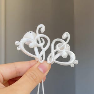 May include: A pair of white earbud covers featuring a decorative treble clef and heart-shaped frame design. Each cover is accented with small, clear, circular rhinestones along the outer edge, providing a detailed musical aesthetic for earbuds.