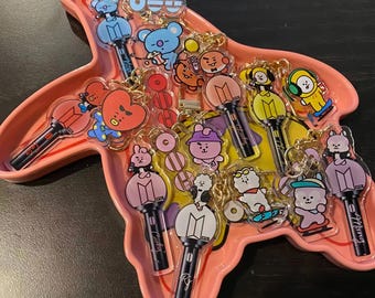 Könnte beinhalten: Eine Sammlung von Acryl-Schlüsselanhängern mit BT21-Figuren und Leuchtstäben, angeordnet auf einem pfirsichfarbenen, walförmigen Tablett. Die Anhänger zeigen verschiedene bunte Zeichentrickfiguren wie Tata, Cooky, RJ und Mang, jeweils kombiniert mit einem Miniatur-Leuchtstab-Design. Das Tablett hat einen glatten, geschwungenen Rand und eine glänzende Oberfläche und enthält mehrere einzelne Anhänger mit goldfarbenen Metallringen.
