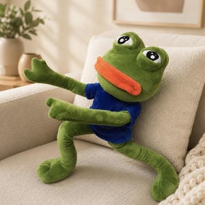 Puede incluir: Un peluche de rana con piel verde, ojos grandes blancos con pupilas negras y labios gruesos de color naranja. La rana lleva una camiseta azul de manga corta y está en una postura dinámica con los brazos extendidos hacia adelante.
