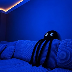 May include: A black plush octopus toy with two large white circular eyes featuring black pupils. The toy has a round head and eight long, dangling tentacles, resting on a blue corduroy sofa under intense blue ambient lighting.