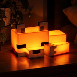 May include: A blocky, pixelated fox-shaped lamp emitting a warm orange glow. The lamp features a rectangular head with dark square ears, eyes, and nose details, and a separate rectangular body section, all constructed from smooth, light-diffusing material.
