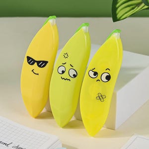 May include: Three yellow banana-shaped erasers with cartoon faces. One has sunglasses, another an angry face, and the third a sad face with a plaster. The erasers are on a white surface.