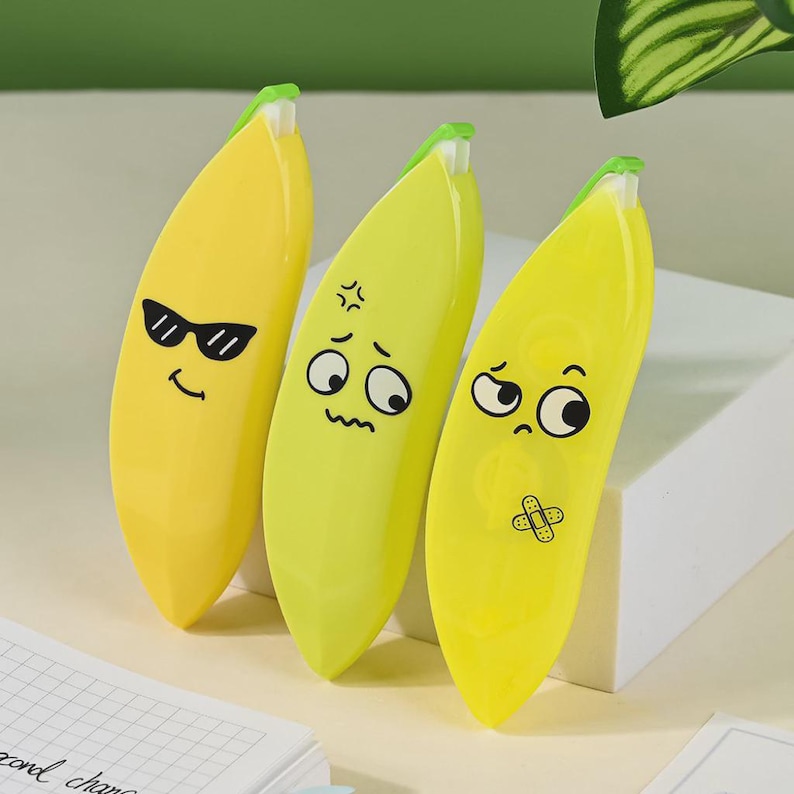 May include: Three yellow banana-shaped erasers with cartoon faces. One has sunglasses, another an angry face, and the third a sad face with a plaster. The erasers are on a white surface.