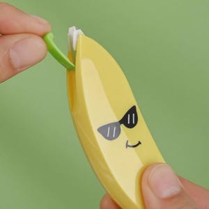 May include: A yellow banana-shaped tape dispenser with a smiling face and sunglasses graphic. A green piece is being removed from the dispenser. The background is a solid green colour.