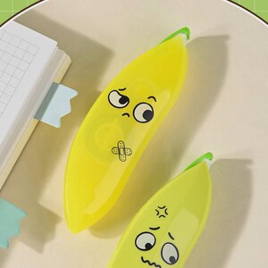 May include: Two yellow banana-shaped tape dispensers with cartoon faces. One has a sad face with a bandage, the other has an angry face. The dispensers have green tips and are on a light-coloured surface.