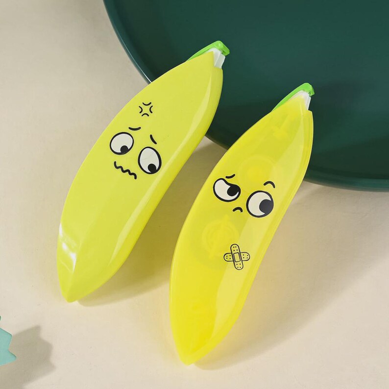 May include: Two yellow banana-shaped correction tape dispensers with cartoon faces. One has an angry expression, the other a sad expression with a bandage graphic. The dispensers have green accents at the tip.