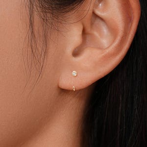 May include: A small gold stud earring featuring a single clear round gemstone set in the center of the earlobe. The earring has a thin, curved gold wire that extends from the back of the stud and loops underneath the bottom of the earlobe.