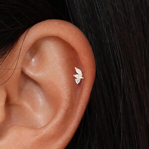 May include: A close-up of an ear with a small, delicate earring. The earring features a cluster of three white gemstones in a leaf or floral design. The earring is set in a gold-colored metal.