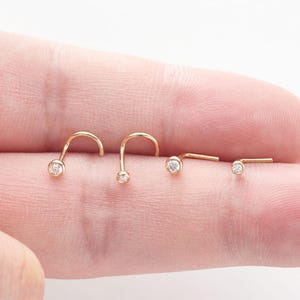 May include: Four gold nose studs, each featuring a small, round, clear gemstone set in a bezel. The studs have a curved L-shaped post design, with the metal showing a polished, warm yellow tone and the stones appearing clear and faceted.