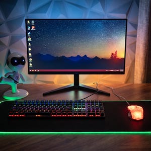 May include: A gaming setup featuring a black monitor displaying a starry night scene. A keyboard and mouse with colorful LED lights sit on a black mousepad with a green illuminated edge. A small robot figurine is on the desk.