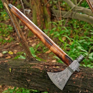 May include: A tomahawk featuring a patterned Damascus steel axe head with a dark, swirling metallic finish and a long, straight wooden handle with a rich, dark brown grain. The axe head is securely attached to the handle with a metal collar.