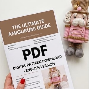 May include: A crochet pattern download titled "The Ultimate Amigurumi Guide" with a pink and tan crocheted nutcracker doll. The digital pattern is in English. The doll has a hat and a jacket with gold trim.