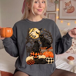 May include: A dark gray crewneck sweatshirt featuring a graphic print of three black cats sitting among various pumpkins and gourds. The design includes a large yellow full moon, bare tree branches, and flying bats against a dark background.