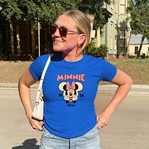 Może przedstawiać: Kr&oacute;lewsko niebieska koszulka z grafiką Myszki Minnie. Grafika zawiera r&oacute;żowy napis "MINNIE", lata "19" i "28" oraz logo Disneya. Koszulka jest z kr&oacute;tkim rękawem.