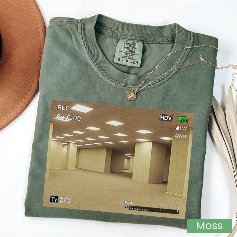 May include: A moss green crew neck t-shirt featuring a graphic print of a yellow-toned, empty office hallway with a digital camera recording interface overlay. The shirt has a Comfort Colors brand label at the inner neckline and a size small tag.