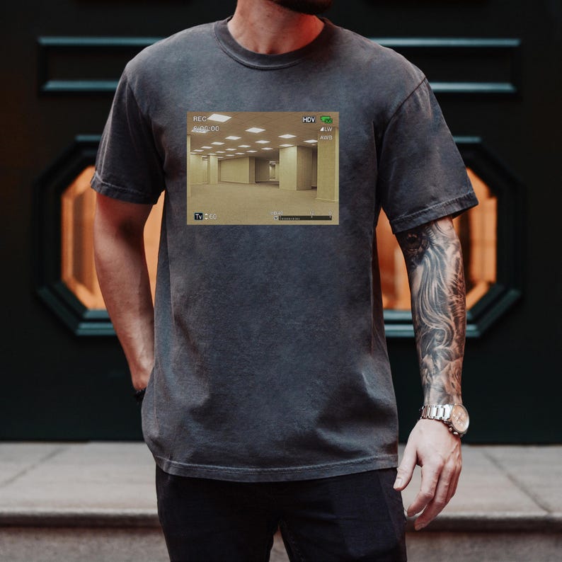 May include: A charcoal gray short-sleeve t-shirt featuring a rectangular graphic print of a yellow-toned, empty office space with fluorescent ceiling lights. The graphic includes a digital camera interface with text reading REC, HDV, and Tv 60.