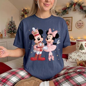 May include: A navy blue t-shirt featuring a graphic of Mickey Mouse dressed as a nutcracker and Minnie Mouse in a pink ballerina outfit, surrounded by small gold stars. The design is centered on the chest of the short-sleeved crew neck shirt.