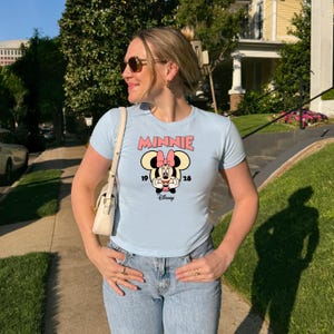 Może przedstawiać: Jasnoniebieska koszulka z grafiką Myszki Minnie. Grafika zawiera r&oacute;żowy napis "MINNIE", wizerunek Myszki Minnie oraz liczby "19" i "28". Koszulka jest zestawiona z niebieskimi dżinsami.