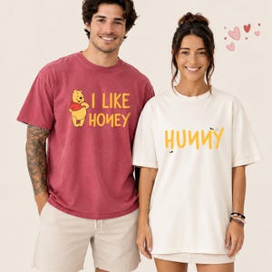 May include: A dark red t-shirt featuring a graphic of Winnie the Pooh and the yellow text I LIKE HONEY, and a cream-colored t-shirt featuring the yellow text HUNNY with small bee illustrations, both with short sleeves and crew necklines.