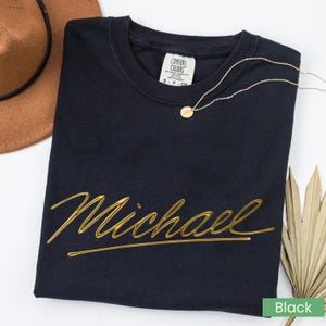 May include: Black t-shirt with the name "Michael" in gold script. A gold necklace with a small pendant rests on the shirt. A brown hat and dried palm leaves are in the background. The shirt has a Comfort Colors tag.