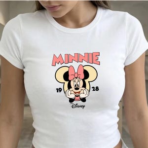 Może przedstawiać: Biała koszulka z grafiką Myszki Minnie. Projekt zawiera r&oacute;żowy napis "MINNIE", kresk&oacute;wkowy wizerunek Myszki Minnie oraz liczby "19" i "28". Obecne jest r&oacute;wnież logo Disneya.