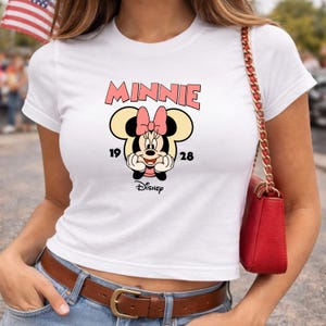Może przedstawiać: Biała koszulka z grafiką Myszki Minnie. Projekt zawiera r&oacute;żowy napis "MINNIE", cyfry "19" i "28" oraz logo Disneya. Grafika przedstawia Myszkę Minnie z r&oacute;żową kokardą.
