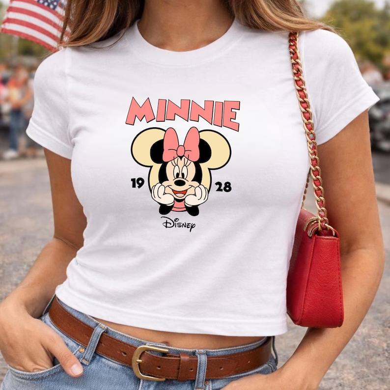 Może przedstawiać: Biała koszulka z grafiką Myszki Minnie. Projekt zawiera r&oacute;żowy napis "MINNIE", cyfry "19" i "28" oraz logo Disneya. Grafika przedstawia Myszkę Minnie z r&oacute;żową kokardą.