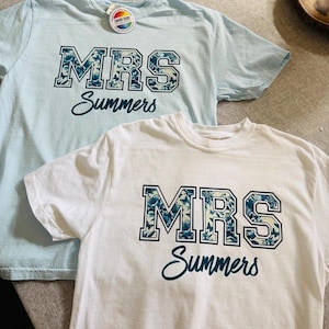 May include: Two short-sleeved t-shirts, one light blue and one white, are displayed. Each shirt features the text "MRS Summers" in a floral design. The light blue shirt has a tag attached. The shirts are likely for a wedding or bridal event.